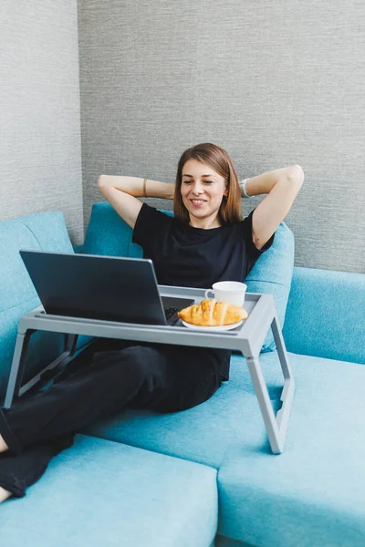 Attractive young woman using laptop computer while sitting on sofa at home with coffee and croissants working. Work online at a distance.