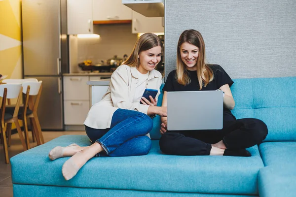 Two beautiful young women using laptop watching movie sitting on sofa at home, two female friends sitting on sofa at home in living room.