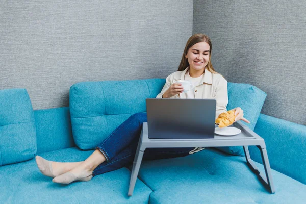 Smiling woman using laptop to check e-mail news online while sitting on sofa, working on computer, writing blog or watching webinar, studying at home