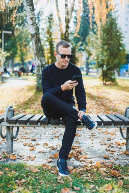 Siyah kazaklı yalnız bir genç adam parkta bir bankta oturur ve telefonda konuşur. Sıcak bir sonbahar günü