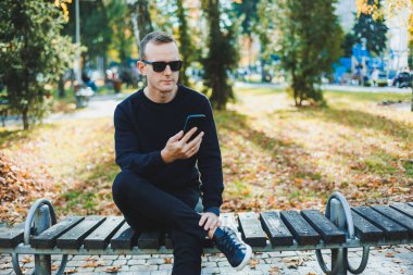 Siyah kazaklı yalnız bir genç adam parkta bir bankta oturur ve telefonda konuşur. Sıcak bir sonbahar günü