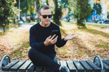 Siyah kazaklı yalnız bir genç adam parkta bir bankta oturur ve telefonda konuşur. Sıcak bir sonbahar günü