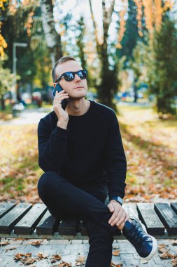 Siyah kazaklı yalnız bir genç adam parkta bir bankta oturur ve telefonda konuşur. Sıcak bir sonbahar günü