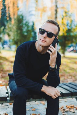 Siyah kazaklı yalnız bir genç adam parkta bir bankta oturur ve telefonda konuşur. Sıcak bir sonbahar günü