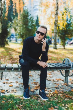 Siyah kazaklı yalnız bir genç adam parkta bir bankta oturur ve telefonda konuşur. Sıcak bir sonbahar günü