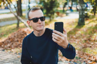 Siyah kazaklı yalnız bir genç adam parkta bir bankta oturur ve telefonda konuşur. Sıcak bir sonbahar günü