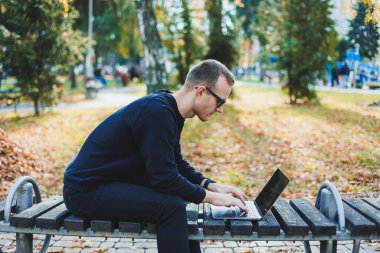 Genç bir adam parkta bir bankta oturuyor ve dizüstü bilgisayarla çalışıyor. Sıcak bir sonbahar günü. Uzak çalışma
