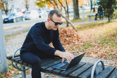 Genç bir adam parkta bir bankta oturuyor ve dizüstü bilgisayarla çalışıyor. Sıcak bir sonbahar günü. Uzak çalışma