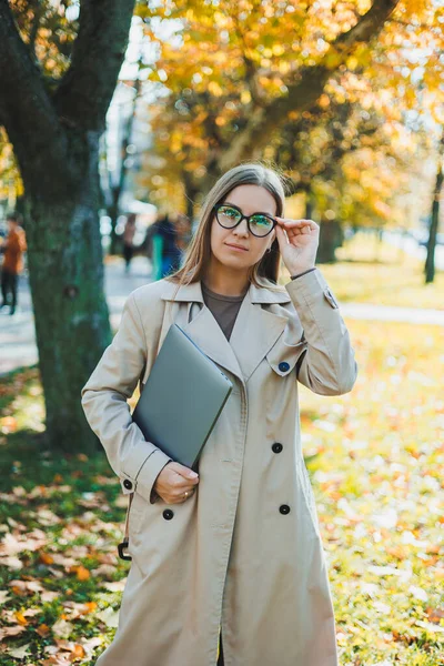 長い髪をした眼鏡をかけた可愛い女性が ノートパソコンを手に秋の公園を歩いている 新鮮な空気中でオンラインで働く — ストック写真