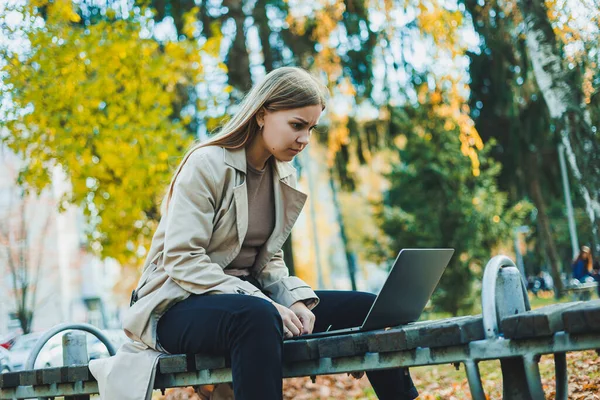 stock image Cute female student sitting in an autumn park with a laptop and working online. Happy woman working outdoors in the park.