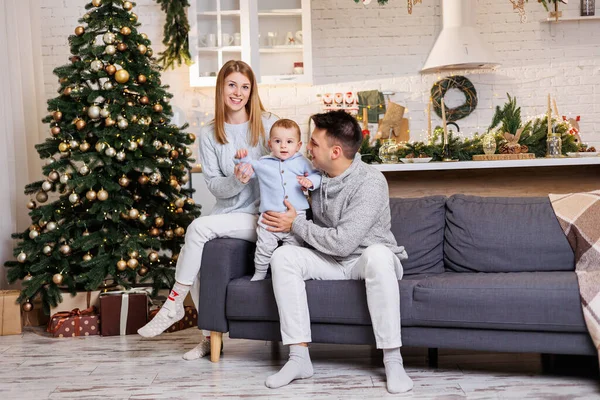 Stock image A happy family is sitting on the couch with their little son near the Christmas tree and smiling joyfully. Home festive atmosphere of the new year.