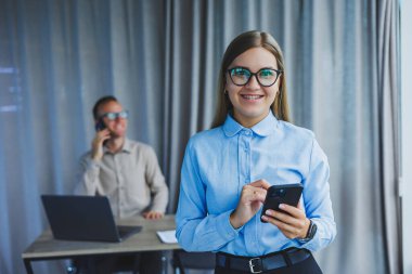 Manager woman in classic glasses smiling during working hours in the office talking on the phone, table with laptop, colleagues in the background. A colleague is in the background, selective focus