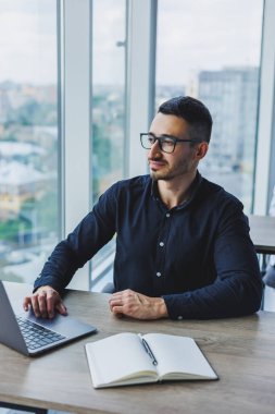 Gözlüklü ve tişörtlü genç başarılı erkek işçi masamızda oturuyor ve modern ofiste dizüstü bilgisayarla ekrana bakarak proje üzerinde çalışıyor.