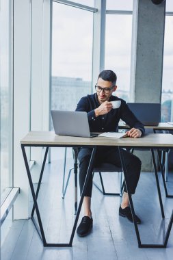 Beyaz gömlekli yakışıklı bir yönetici bir masada oturuyor ve modern bir ofiste kahve içiyor. Ofiste çalışma ortamı.