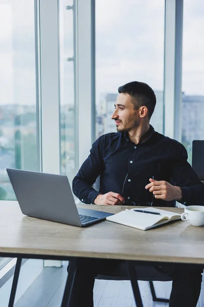 一位身穿黑色衬衫 面带微笑的商人正坐在办公桌前使用笔记本电脑 一个人在办公室的电脑上工作 分析数据 网络营销 成人教育 电子学习 — 图库照片