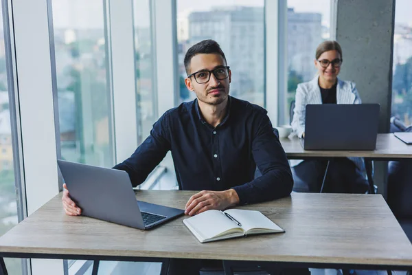 stock image Attractive business man of Caucasian appearance in a black shirt sits at a desk using a laptop in a modern office with colleagues in the background. Working atmosphere in the office.