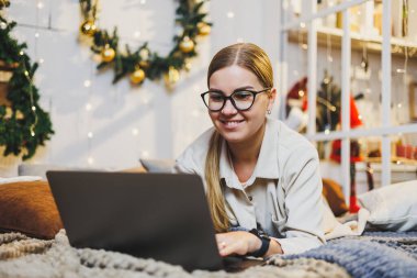 Konforlu ev kıyafetleri içinde genç bir kadın Noel ağacıyla aynı odada dizüstü bilgisayar kullanıyor. Noel 'de evde tasasız, mutlu bir kızın portresi.
