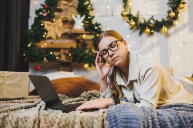 Konforlu ev kıyafetleri içinde genç bir kadın Noel ağacıyla aynı odada dizüstü bilgisayar kullanıyor. Noel 'de evde tasasız, mutlu bir kızın portresi.