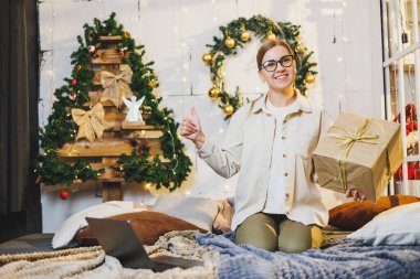 Noel 'e hazırlanmak için dizüstü bilgisayar kullanan sevimli genç bir kadın. Evde Noel ağacının yanında otururken internetten hediye arıyor. Yeni Yıl