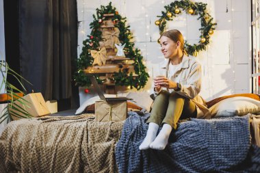 Sarışın, uzun saçlı, elinde kahveyle dizüstü bilgisayar kullanarak evde Noel ağacıyla yatakta gülümseyen genç ve güzel bir kadın. Yeni yıl tatili.