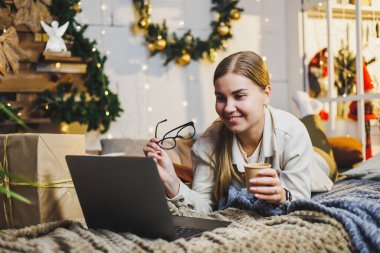 Sarı saçlı, rahat elbiseli, elinde kahve olan, uzaktan kumandalı bilgisayarını kullanan ve evde Noel ağacıyla yatakta gülümseyen güzel bir kadın. Yeni yıl tatili.