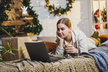 Sarı saçlı, rahat elbiseli, elinde kahve olan, uzaktan kumandalı bilgisayarını kullanan ve evde Noel ağacıyla yatakta gülümseyen güzel bir kadın. Yeni yıl tatili.