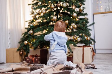 2 yaşında küçük bir çocuk oyuncaklarla süslenmiş bir Noel ağacının yanında oturuyor. Yeni yıl atmosferi