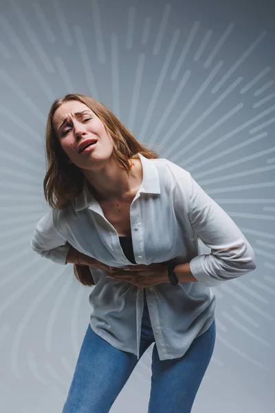 stock image Young smiling woman in a white shirt, black t-shirt and jeans on a gray background. Portrait of a brunette woman she has a pain in the liver, emotion of pain on her face