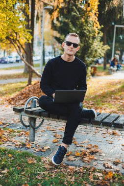 Siyah kazaklı ve güneş gözlüklü çekici bir genç, sonbahar parkında bir bankta oturuyor ve dizüstü bilgisayarla çalışıyor.