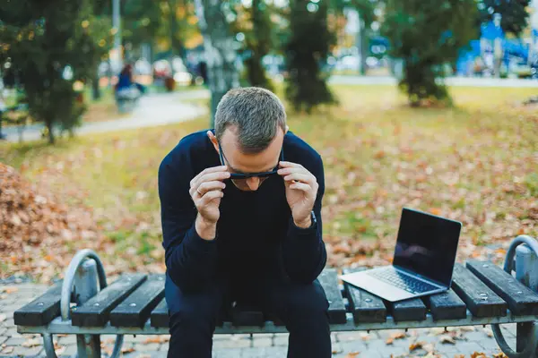 Güneş gözlüklü yakışıklı genç adam, güneşli bir günde parktaki bankta dizüstü bilgisayar ve telefonla oturuyor ve başka tarafa bakıyor. Serbest çalışma, uzak çalışma kavramı.