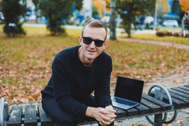 Güneş gözlüklü yakışıklı genç adam, güneşli bir günde parktaki bankta dizüstü bilgisayar ve telefonla oturuyor ve başka tarafa bakıyor. Serbest çalışma, uzak çalışma kavramı.