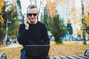 Güneş gözlüklü yakışıklı genç adam, güneşli bir günde parktaki bankta dizüstü bilgisayar ve telefonla oturuyor ve başka tarafa bakıyor. Serbest çalışma, uzak çalışma kavramı.