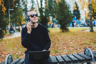 Güneş gözlüklü yakışıklı genç adam, güneşli bir günde parktaki bankta dizüstü bilgisayar ve telefonla oturuyor ve başka tarafa bakıyor. Serbest çalışma, uzak çalışma kavramı.