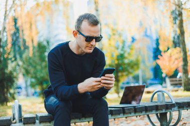 Güneş gözlüklü yakışıklı genç adam, güneşli bir günde parktaki bankta dizüstü bilgisayar ve telefonla oturuyor ve başka tarafa bakıyor. Serbest çalışma, uzak çalışma kavramı.