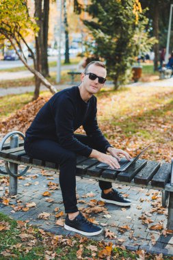 Genç bir adam parkta bir bankta oturuyor ve dizüstü bilgisayarla çalışıyor. Sıcak bir sonbahar günü. Uzak çalışma