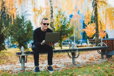 Siyah kazaklı ve güneş gözlüklü çekici bir genç, sonbahar parkında bir bankta oturuyor ve dizüstü bilgisayarla çalışıyor.