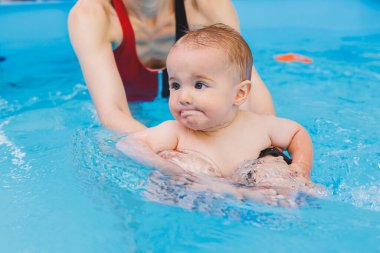 Güzel bir anne küçük oğluna havuzda yüzmeyi öğretir. Bir çocuk annesiyle birlikte suda eğleniyor. Çocuk gelişimi. Çocuklar için ilk yüzme dersleri