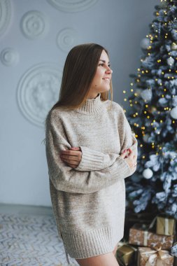 Süslü bir süveter giymiş, yüzünde bir gülümsemeyle genç, sarışın bir kadın süslü bir Noel ağacının yanında duruyor. Yeni yılın kutlaması..