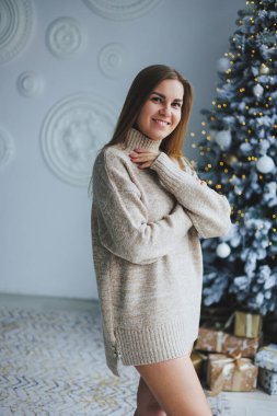 Süslü bir süveter giymiş, yüzünde bir gülümsemeyle genç, sarışın bir kadın süslü bir Noel ağacının yanında duruyor. Yeni yılın kutlaması..