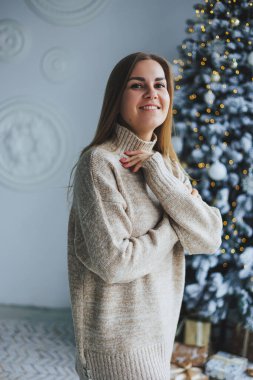 Süslü bir süveter giymiş, yüzünde bir gülümsemeyle genç, sarışın bir kadın süslü bir Noel ağacının yanında duruyor. Yeni yılın kutlaması..