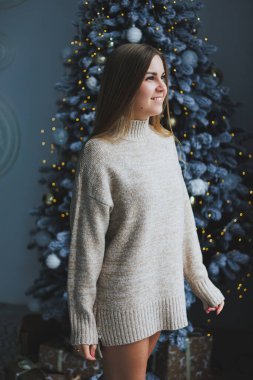 Süslü bir süveter giymiş, yüzünde bir gülümsemeyle genç, sarışın bir kadın süslü bir Noel ağacının yanında duruyor. Yeni yılın kutlaması..