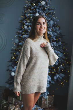 Süslü bir süveter giymiş, yüzünde bir gülümsemeyle genç, sarışın bir kadın süslü bir Noel ağacının yanında duruyor. Yeni yılın kutlaması..