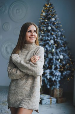 Süslü bir süveter giymiş, yüzünde bir gülümsemeyle genç, sarışın bir kadın süslü bir Noel ağacının yanında duruyor. Yeni yılın kutlaması..