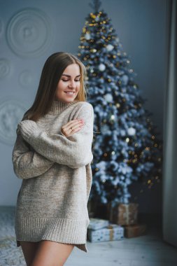 Süslü bir süveter giymiş, yüzünde bir gülümsemeyle genç, sarışın bir kadın süslü bir Noel ağacının yanında duruyor. Yeni yılın kutlaması..