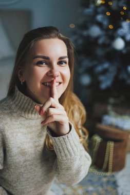 Süslü bir süveter giymiş, yüzünde bir gülümsemeyle genç, sarışın bir kadın süslü bir Noel ağacının yanında duruyor. Yeni yılın kutlaması..