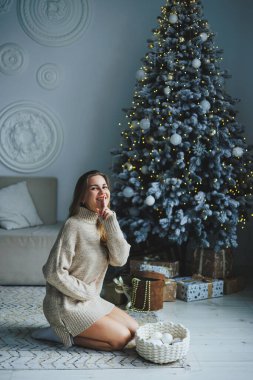 Süslü bir süveter giymiş, yüzünde bir gülümsemeyle genç, sarışın bir kadın süslü bir Noel ağacının yanında duruyor. Yeni yılın kutlaması..