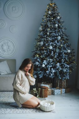 Süslü bir süveter giymiş, yüzünde bir gülümsemeyle genç, sarışın bir kadın süslü bir Noel ağacının yanında duruyor. Yeni yılın kutlaması..