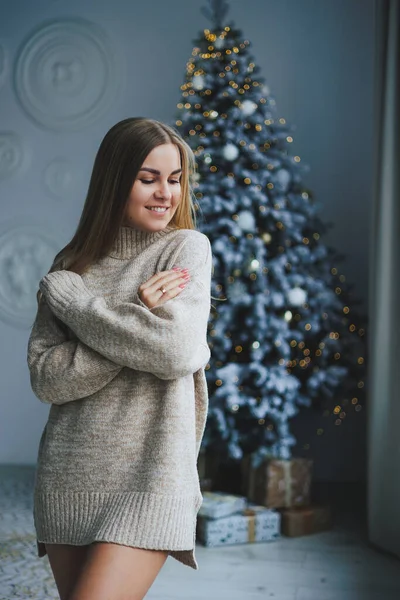 Süslü bir süveter giymiş, yüzünde bir gülümsemeyle genç, sarışın bir kadın süslü bir Noel ağacının yanında duruyor. Yeni yılın kutlaması..