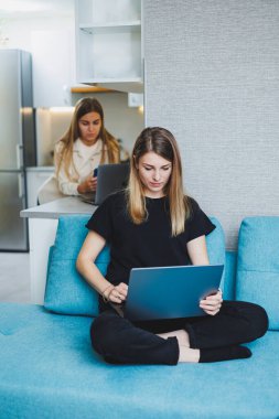 Genç ve güzel bir kadın evdeki kanepede oturuyor ve dizüstü bilgisayarla çalışıyor, arka planda başka bir kadın mutfakta telefonla konuşuyor. İki arkadaşım evde çalışıyor..