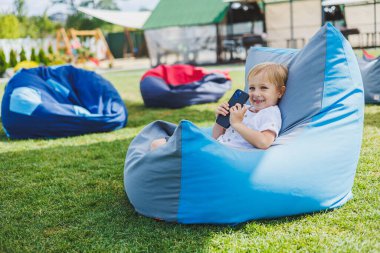 5 yaşında küçük, sarışın bir çocuk sokakta bir sandalyede oturur ve telefona bakar. Elinde aleti olan modern bir çocuk.